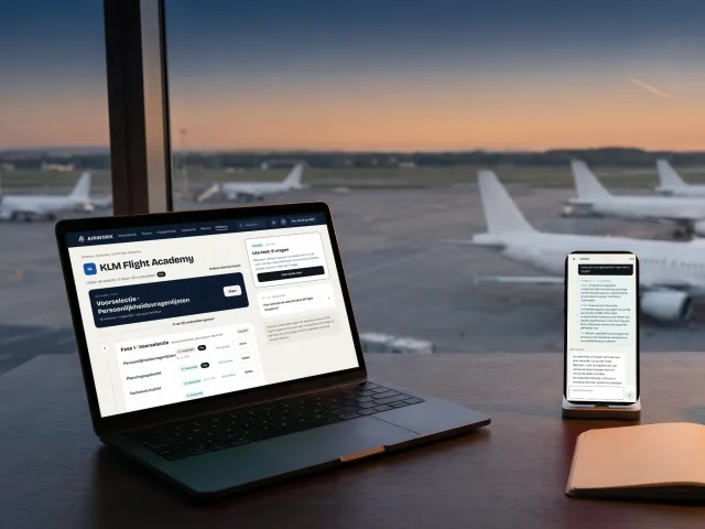 Een oefenroute voor een pilotenselectie op Airwork op een laptop, en de interview-simulator op een telefoon, aan een raam met uitzicht op een luchthaven
