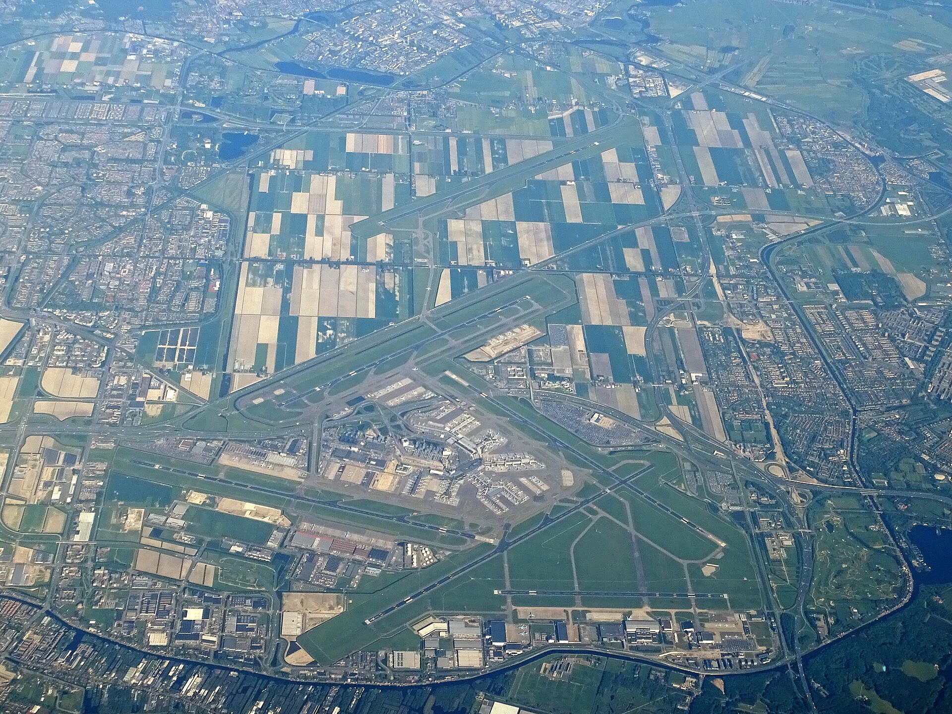Luchtfoto van Schiphol met de start- en landingsbanen, de terminal en de omliggende akkers en woonwijken
