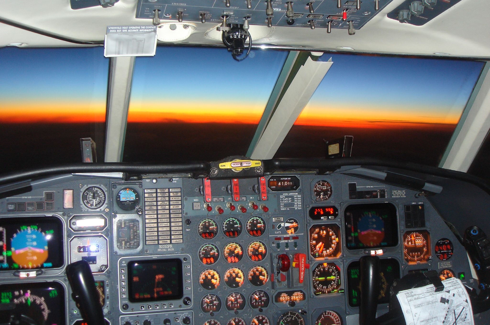 Zicht vanuit een cockpit bij zonsopkomst na een nachtvlucht: een oranje horizon boven een donkere aarde en een verlicht instrumentenpaneel