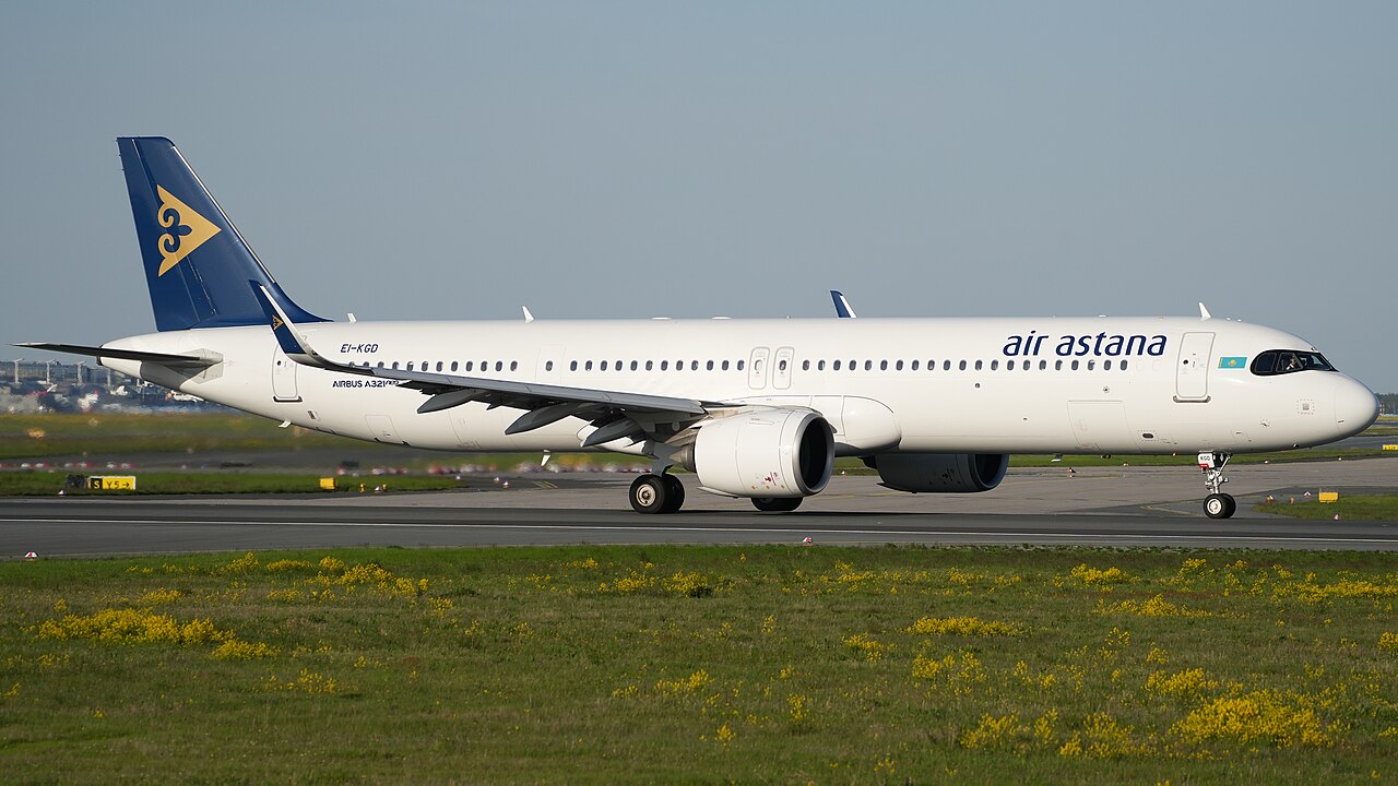 Een wit Airbus A321neo-verkeersvliegtuig van Air Astana met donkerblauwe staart en gouden logo op de startbaan, met gras en gele bloemen op de voorgrond
