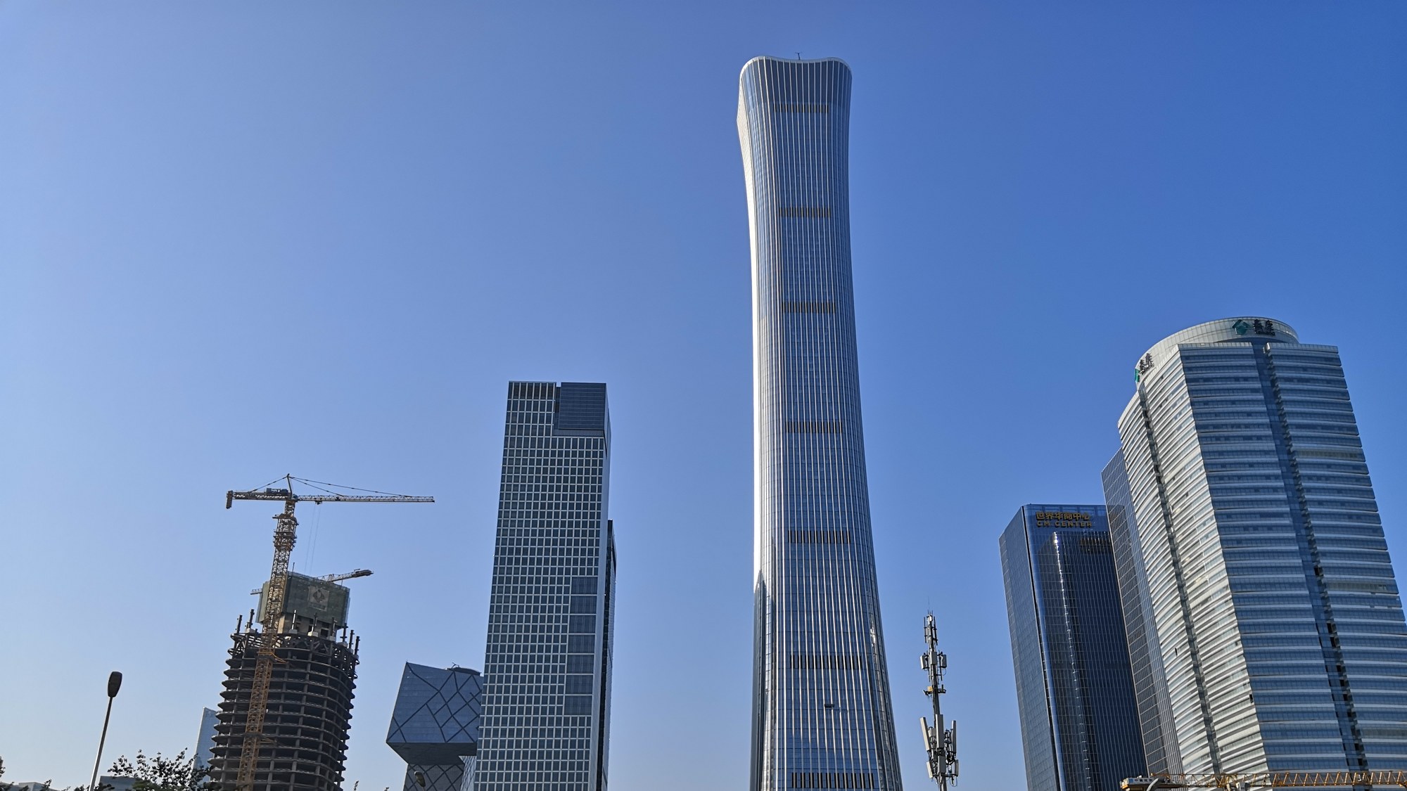 De slanke, naar boven toe uitwaaierende CITIC Tower in het zakendistrict van Peking tegen een strakblauwe lucht, tussen lagere kantoortorens en een bouwkraan