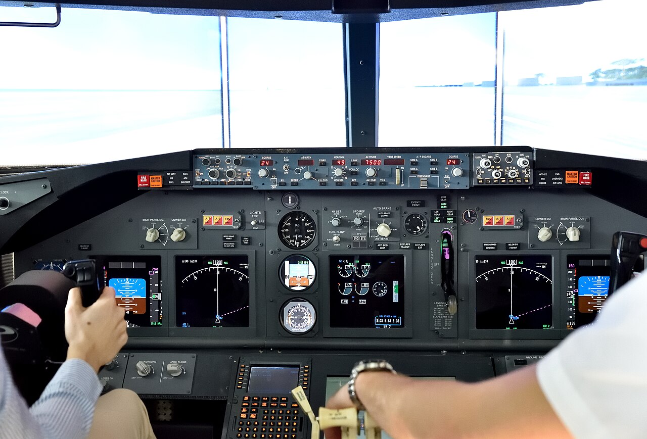 Twee mensen aan de bediening in de cockpit van een Boeing 737-800-simulator, met de instrumentenpanelen en het projectiescherm voor het zicht naar buiten