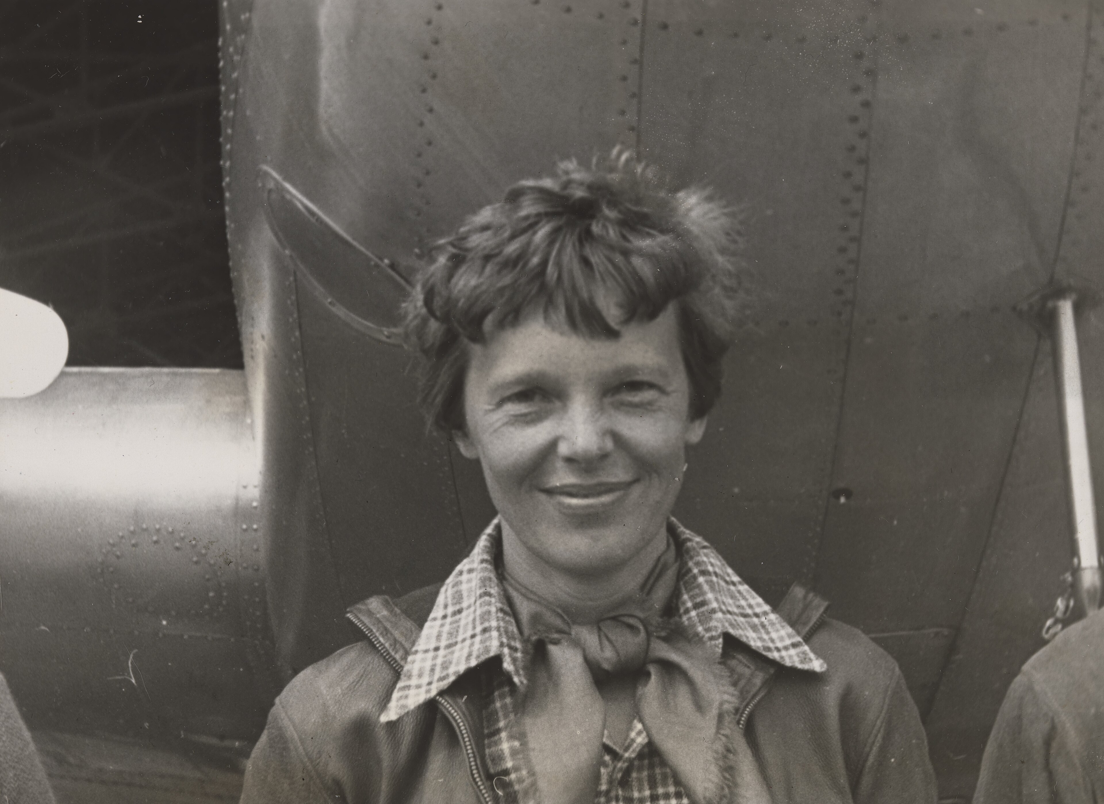 Zwart-witfoto van Amelia Earhart met kort krullend haar, geruit hemd en leren jack, glimlachend onder de neus van haar Lockheed Electra