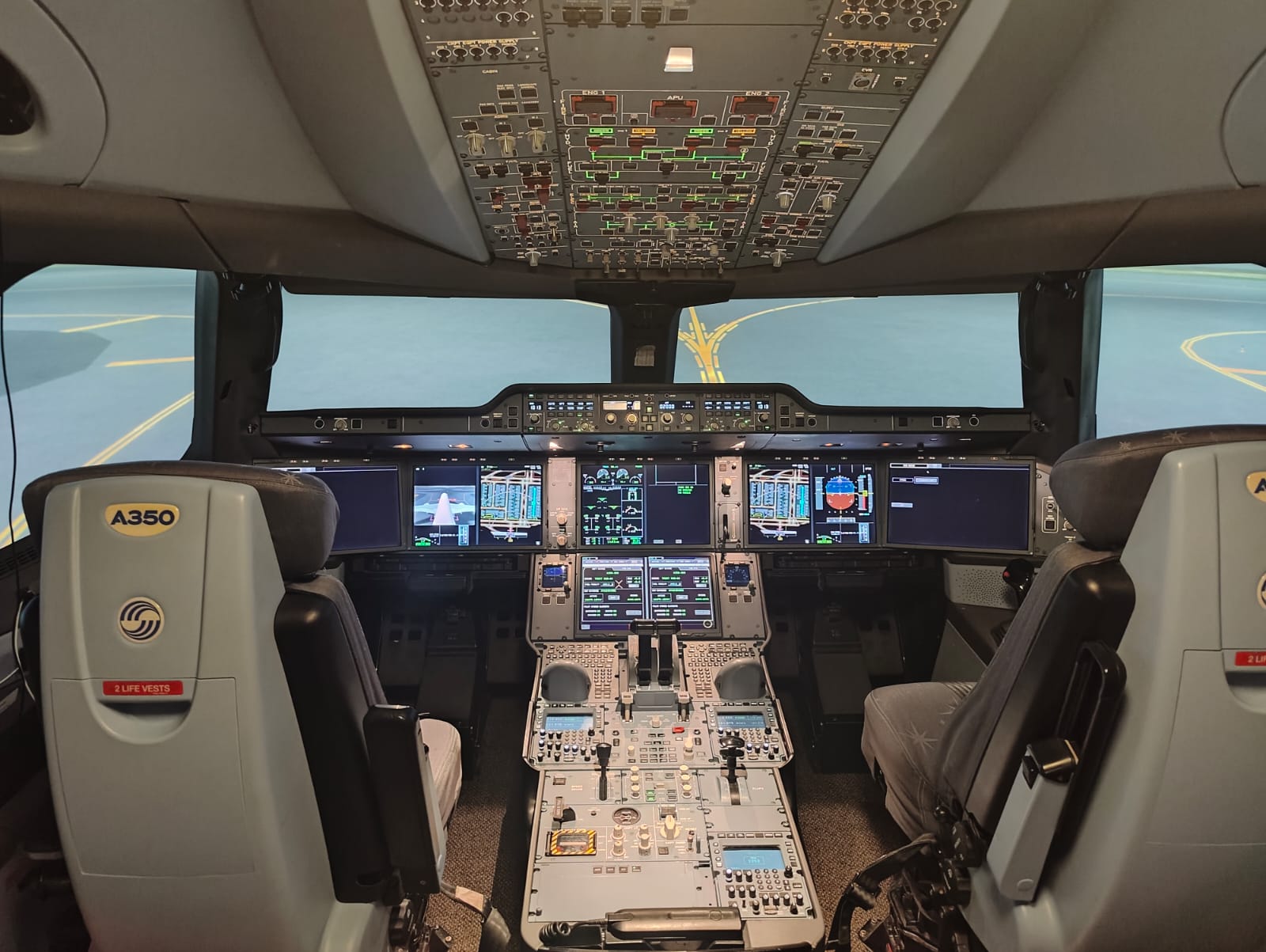 Cockpit van een full flight simulator van de Airbus A350-900 met twee lege pilotenstoelen, de schermen aan en op het buitenzicht de gele lijnen van een taxibaan