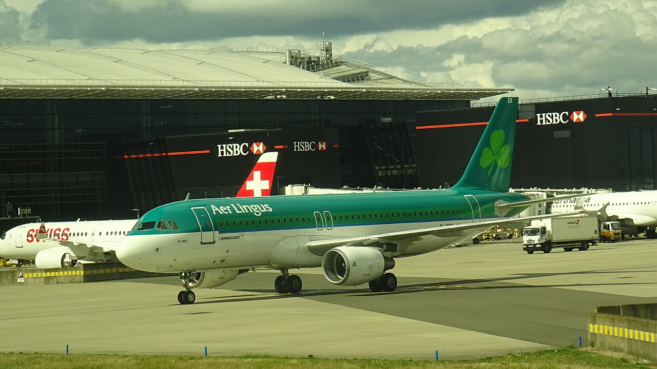 Een Airbus A320 van Aer Lingus met groene staart en klavertje taxiet op London Heathrow, met op de achtergrond een terminal en een toestel van Swiss